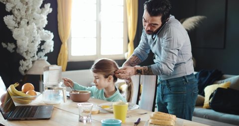 Business dad make  hairstyle to  his daughter. Businessman is preparing his daughter for school and talking on the phone. Family morning over breakfast. Getting ready for work and school