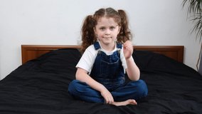 Happy little girl smiling looking at camera sit on cozy bed at home, funny kid talking to webcam making online video call or recording vlog having fun, preschool child with pretty face waving hand 
