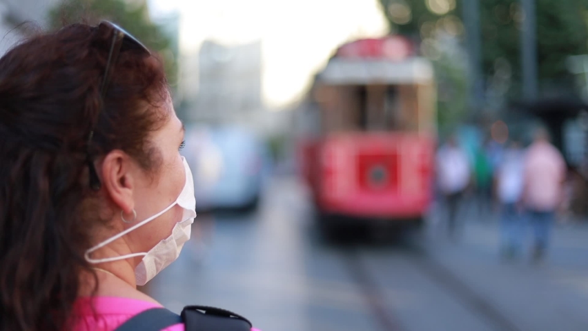 A brunette woman in a protective mask on her face walks along the city streets and looks at the sights, a red retro tram is passing by.