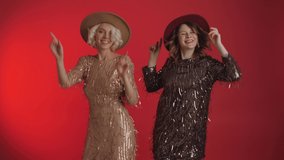 Joyful women wearing sequin dresses are dancing at the party. Out Of Hat falling confetti. Celebrating New year, Christmas, birthday concept. Isolated on red background.