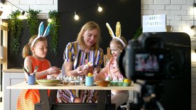 Mom blogger is decorating Easter eggs with her daughters at home indoors. Family records the process of panting eggs on camera. Happy Easter holidays. 