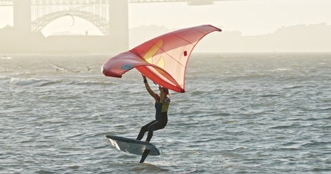 San Francisco October 2020. Slow motion strong athletic kite-surfer female riding with world famous Golden Gate Bridge on background at sunset. Woman on airush freewing at strong wind in stormy waters