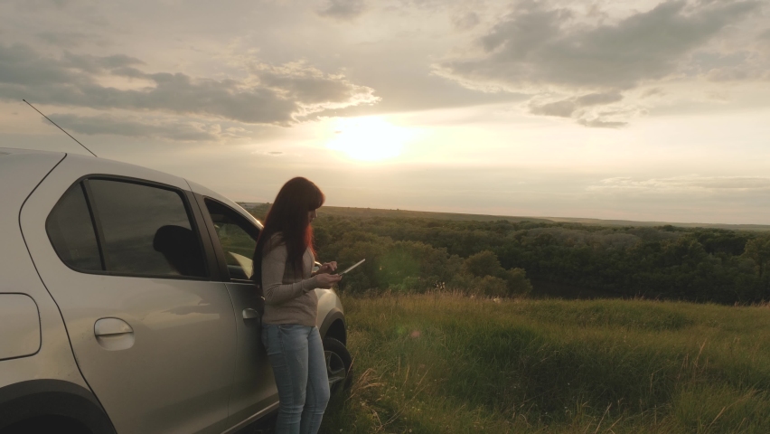 A female tourist driver with tablet stopped in camping car, looking at sunset from mountain. Free woman, traveling by car, checking route in navigator. Driver holding modern gadget in hands, rest.