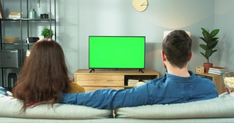 Rear of Caucasian married family young couple watching TV with chroma key and eating popcorn while sitting at home on sofa. Man and woman watch film on television in room, leisure concept