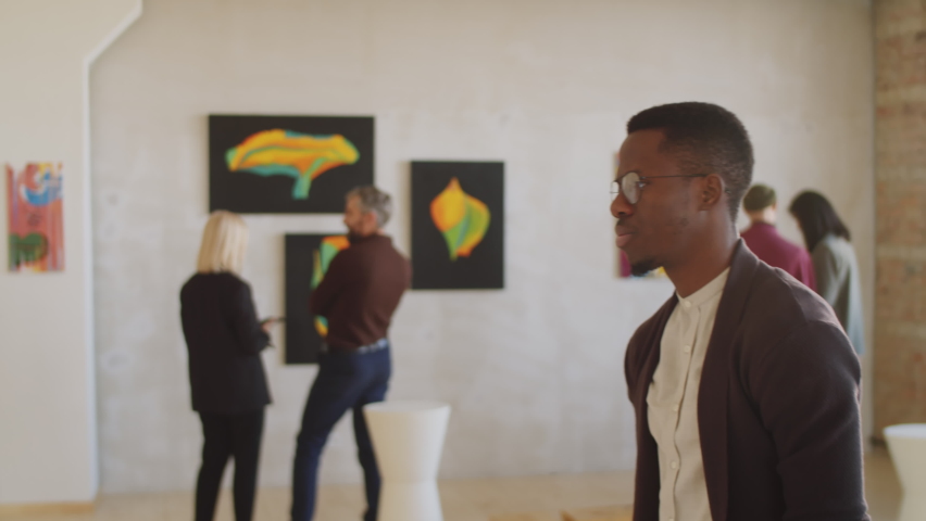 Side view of young Afro-American man rubbing his chin and looking at painting on the wall pensively while visiting modern art exhibition in gallery