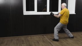 An old male athlete with black pads on his arms trains two punches with a half-jump