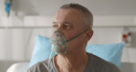 Portrait of elderly person with oxygen mask sitting up in hospital bed and looking around. Sick aged male patient breathing in oxygen mask sitting in bed at hospital ward