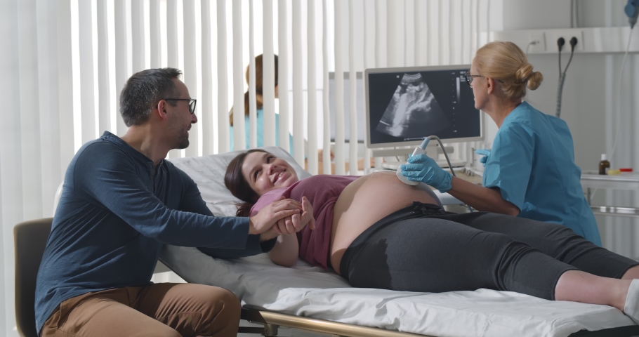 Happy couple making ultrasound of baby in clinic office. Senior doctor gynecologist performing screening of pregnant woman visiting hospital with supporting husband | Shutterstock HD Video #1069624237