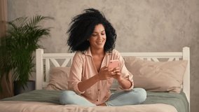 Portrait cute African American woman talking on a video call using mobile phone. Black female posing sitting on bed in a bedroom with bright room interior. Slow motion ready, 4K at 59.97fps.