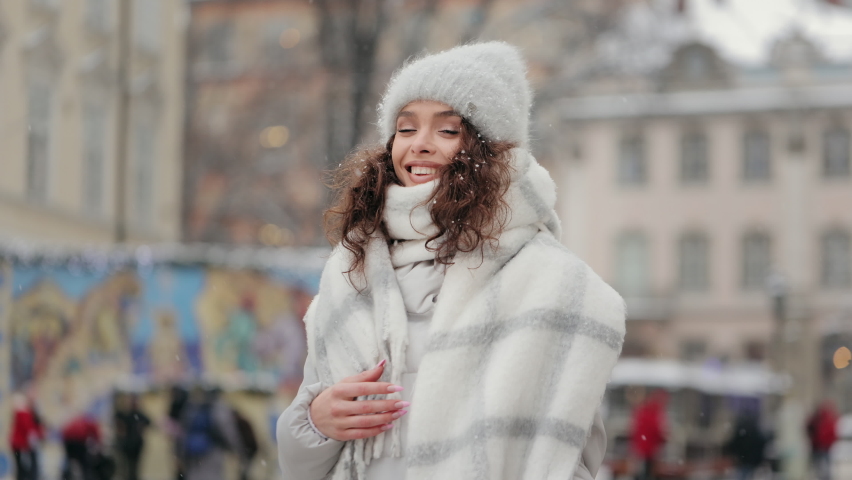 A woman is standing in the center of the city and smiling. She is looking at the camera and holding shopping bags on her shoulder. Snow is falling. 4K