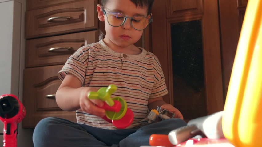 Caucasian small boy with glasses sitting on the floor and trying to fix a constructor toy