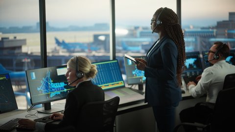 Black Female Air Traffic Controller Holding Tablet in Airport Tower. Office Room is Full of Desktop Computer Displays with Navigation Screens, Airplane Departure and Arrival Data for the Team.