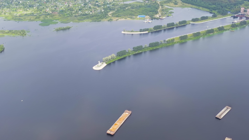 Rybinsk, Russia. The Mother Volga monument stands on a small island near the shipping locks of the Rybinsk Reservoir, visible as a bright white sculptural figure rising above the water surface. The aerial perspective captures the full length of the lock system cutting across the wide reservoir, with tree-lined embankments separating the calm water channels. Several empty barges rest on the dark water in the foreground. The surrounding landscape shows a mix of dense green forest and scattered residential development under soft, overcast summer light. The monument was unveiled in 1953 by sculptors Sergei Shaposhnikov and Vera Malashkina, and its reinforced concrete figure reaches a total height of 28 meters including the granite pedestal, making it a distinctive landmark for vessels passing through the Volga-Baltic Waterway.