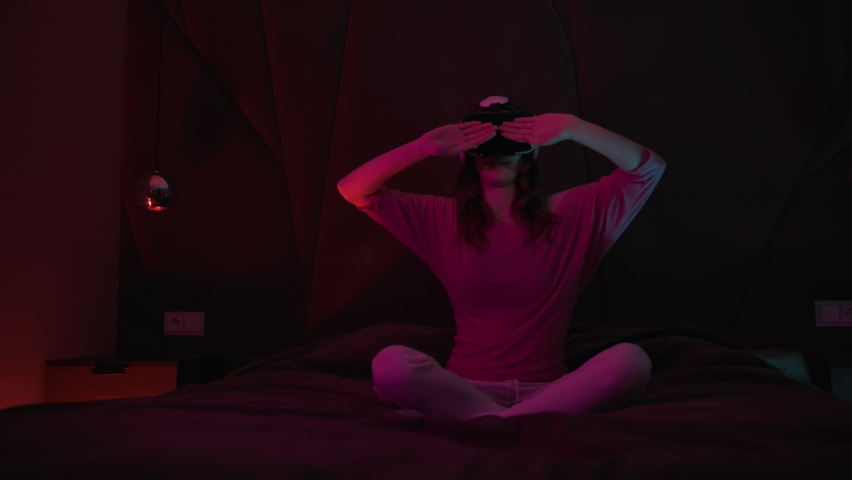 Young Woman Uses Virtual or Augmented Reality Glasses Sitting On A Bed in Dark Room, Female VR Headset User On Digital Interactive Art Performance, Entertainment Of Future Royalty-Free Stock Footage #1069636192