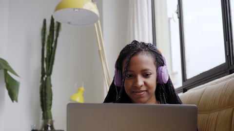 Smiling young woman sitting on sofa with headphones working from home with laptop. Vídeo Stock