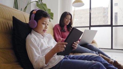 Boy with headphones playing with a tablet on the sofa at home while his mother works remotely on her laptop. Concept of remote work and family reconciliation. Vídeo Stock
