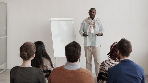 Medium long shot of young African male office worker standing in front of unrecognizable audience, talking, making business presentation Video de stock