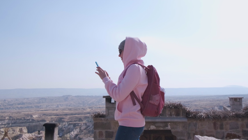 Woman With Red Backpack and Pink Hoodie. Tourist. Smartphone In Hands. Takes Photo Of Stunning View. Stands on Top of Mountain. Ancient Place. Old Town. Vacation Trip. Keepsake Photo. Girl on Vacation