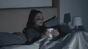 Night home leisure. Late movie. Bedtime entertainment. Relaxed woman with gold eye patches enjoying watching comedy TV show drinking tea alone in cozy bed in dark bedroom with flicker blue light.