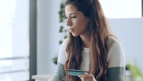 Video of cute young woman holding white credit card for shopping online with computer while sitting in the kitchen at home.