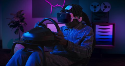 A guy is playing racing game in VR. He is sitting on the gaming chair with a steering wheel and pedals. A guy wears VR headset. Chair is located in the living room with red and blue neon lighting. 