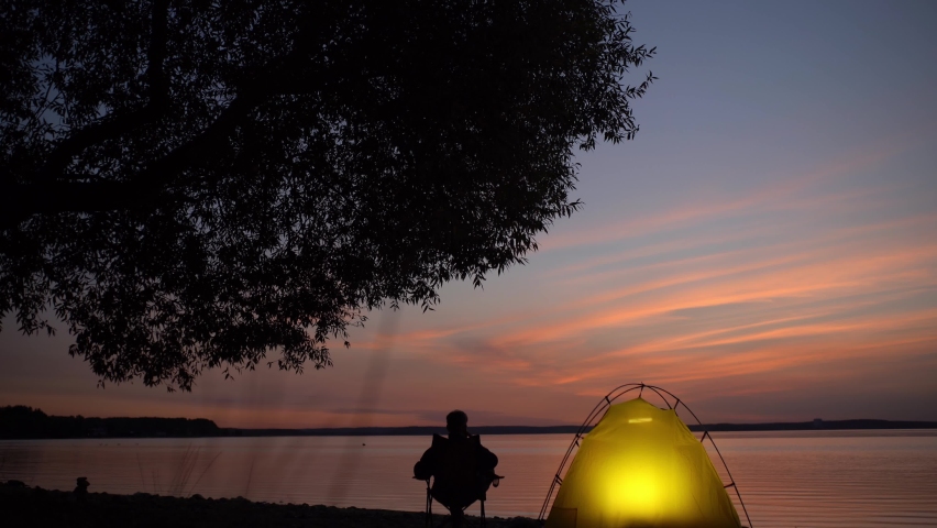 The traveler sits on a camping chair near the glowing tent 
