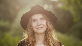 Closeup view video portrait of beautiful young woman wearing black hat and coat stands outdoor in green sunset scenic landscape. Woman looks at camera smiling calmly