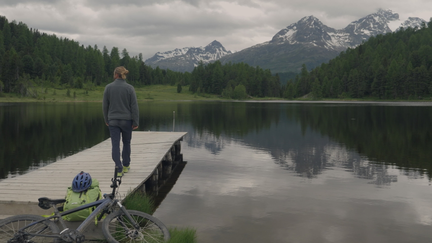 Young man walks on wooden pier above alpine lake. Male on vacation contemplating nature 