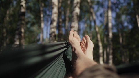 Male feet swinging in green hammock under birch trees in the sun. Slow motion