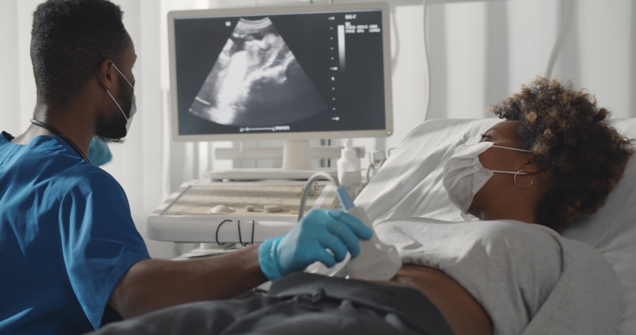 Competent afro male doctor in medical protective mask doing ultrasound screening for pregnant woman at modern clinic. Young afro woman in safety mask visiting gynecologist Royalty-Free Stock Footage #1069670203