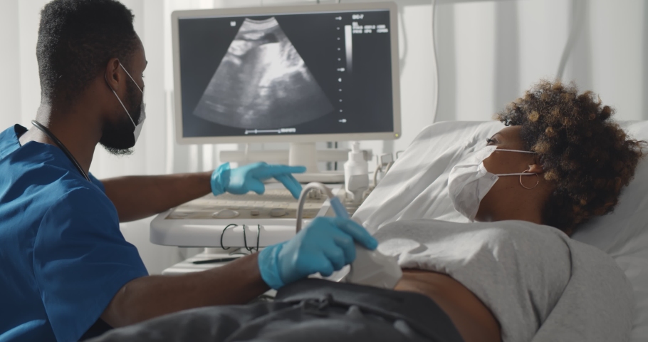 Competent afro male doctor in medical protective mask doing ultrasound screening for pregnant woman at modern clinic. Young afro woman in safety mask visiting gynecologist | Shutterstock HD Video #1069670203