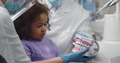 Dentist teaching cute afro-american little girl how to brush teeth. Close up of stomatologist showing child patient how to brush teeth using tooth-brushing on model