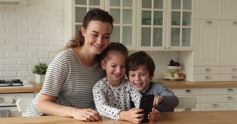 In cozy kitchen young woman and her little children holding smartphone using mobile applications, look at cellphone screen making selfie photo. Family having fun, modern wireless tech usage concept