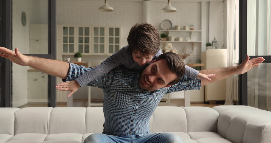 Happy cheerful father sit on couch play with lovely preschool son, spread arms like airplane wings piggybacking carrying on back cute little funny kid boy. Have fun at home, playtime, leisure concept Royalty-Free Stock Footage #1069685578