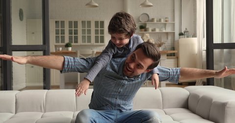 Happy cheerful father sit on couch play with lovely preschool son, spread arms like airplane wings piggybacking carrying on back cute little funny kid boy. Have fun at home, playtime, leisure concept