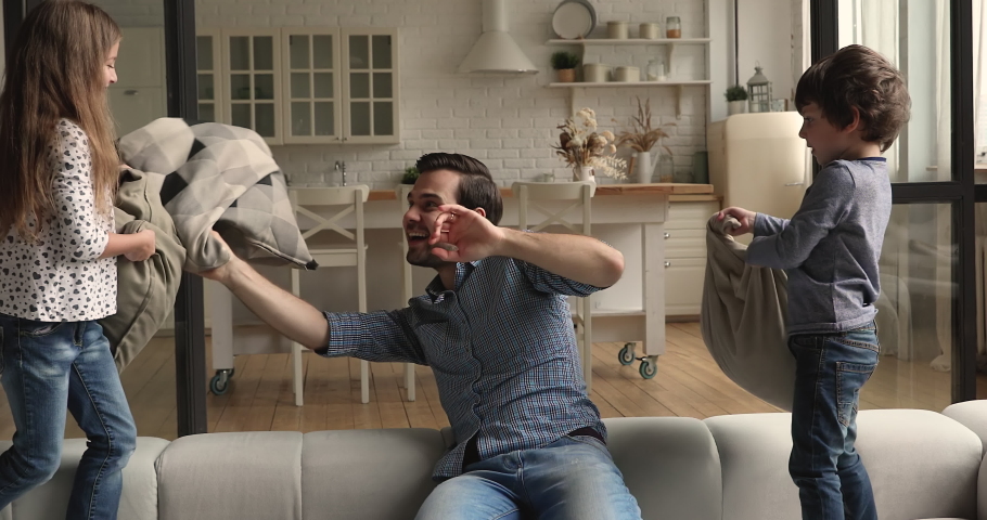 Man plays pillow fight with little son and daughter on sofa. Happy family spend active weekend time at quarantine stay at home enjoy funny playtime holding cushions beating laughing having fun concept
