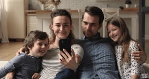 Couple and children sit on sofa having fun using smartphone, enjoy new cool mobile funny app, making video call communication to friends remotely. Family and modern tech online amusement usage concept