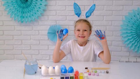 KRASNODAR, RUSSIA - 19 March , 2021. A child with bunny ears holds an Easter egg in his hand. Little girl waving hand painted paint. Easter. Home decoration in blue tones. Slow motion 