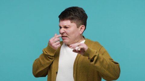 Confused shocked middle aged dark haired man grabbing nose with fingers feeling disgusted unpleasant smell of dirt, harmful emissions. Indoor studio shot isolated on blue background