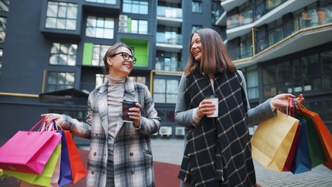 Two happy women walk with shopping bags and takeaway coffee after a successful shopping and talk with interest among themselves. Slow motion