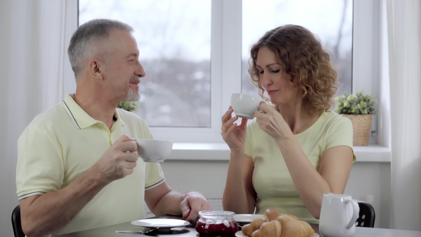 A beautiful middle-aged couple is having breakfast, drinking coffee and talking at the kitchen table sitting at the window of their house.