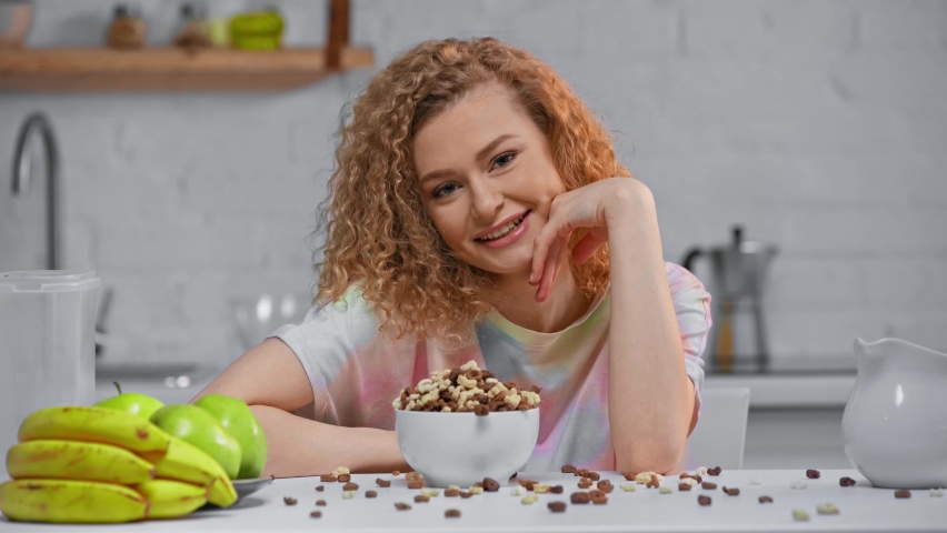 Smiling woman eating cereals near fruits on table