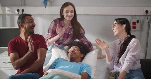Portrait of diverse young people visiting sick african man in hospital laughing and waving hands at camera. Happy afro male patient in hospital bed smiling and talking to friends visitors