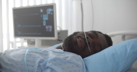 African dead man with oxygen nasal tube lies in hospital bed. Portrait of afro-american patient in coma dying in hospital ward with zero heart rate on monitor