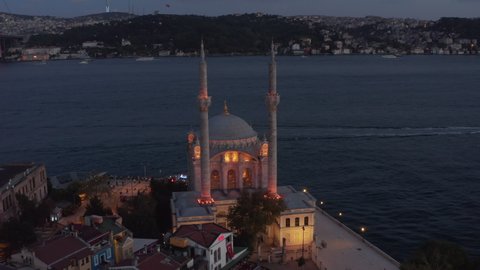 Ortakoy Mosque illuminated in beautiful yellow light on Water side at Dusk with Bosphorus and Bridge, Aerial wide shot