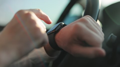 Close-up male hands using modern smart watch in car. Man watch notification. Male hand with a smartwatch holds the steering wheel. Businessman driving car at sunset