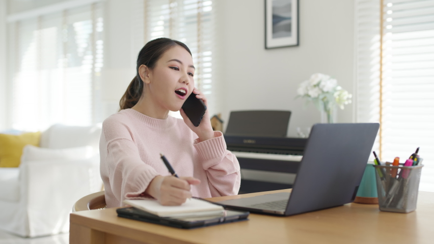 Young happy attractive beautiful asia female staff laugh talk on mobile phone relax comfort sit work on desk table at home. Remotely work on distance job telesale or secretary career for business. | Shutterstock HD Video #1069728361