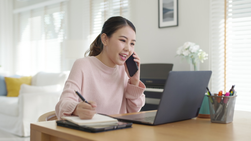 Young happy attractive beautiful asia female staff laugh talk on mobile phone relax comfort sit work on desk table at home. Remotely work on distance job telesale or secretary career for business. | Shutterstock HD Video #1069728361