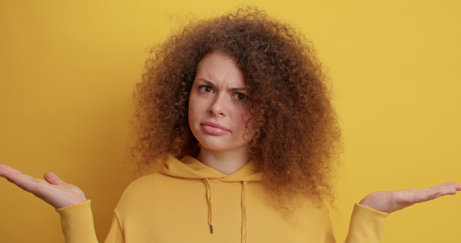 Lovely curly haired young woman raises palms shrugs shoulders feels doubt dressed in hoodie cannot make choice isolated over yellow background. Clueless European female face difficult choice Royalty-Free Stock Footage #1069732060