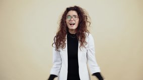 The surprised, victorious young woman with curly hair is surprised and says wow, waving her hands with her fingers in her fist. Young hipster in gray jacket and white shirt, with glasses posing
